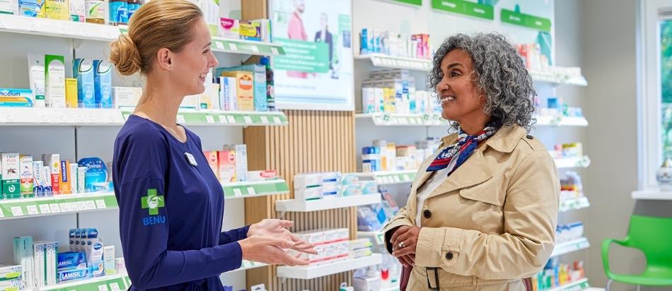 Twee vrouwen in gesprek in een apotheek, omringd door schappen met medicijnen en gezondheidsproducten.