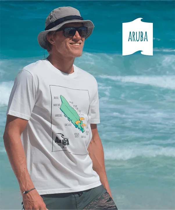 Young man stands smiling on the beach wearing a white Swakiko T-shirt with the color design of Aruba depicted as damaged surfboard