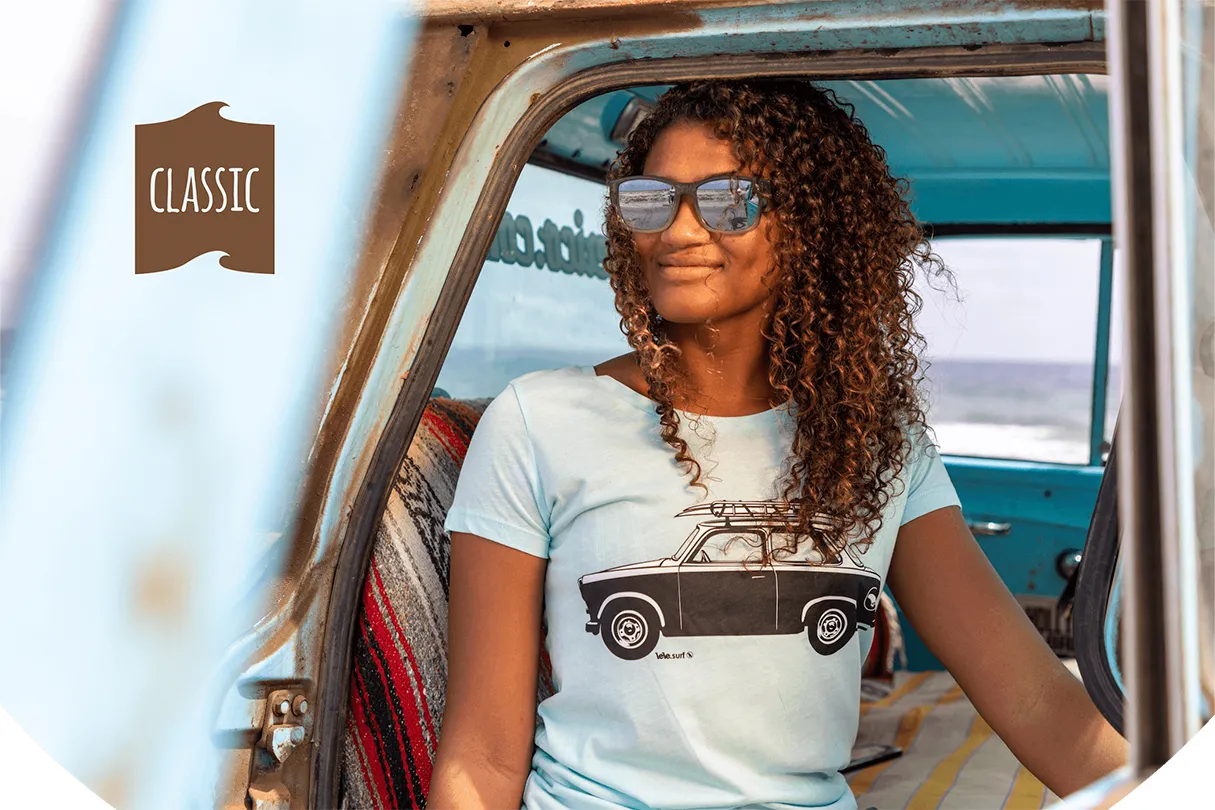 Young lady sits in a Cadillac looking out in front of her through sunglasses and wearing a turquoise Swakiko T-shirt with the print of a Trabant with surfboards on the roof