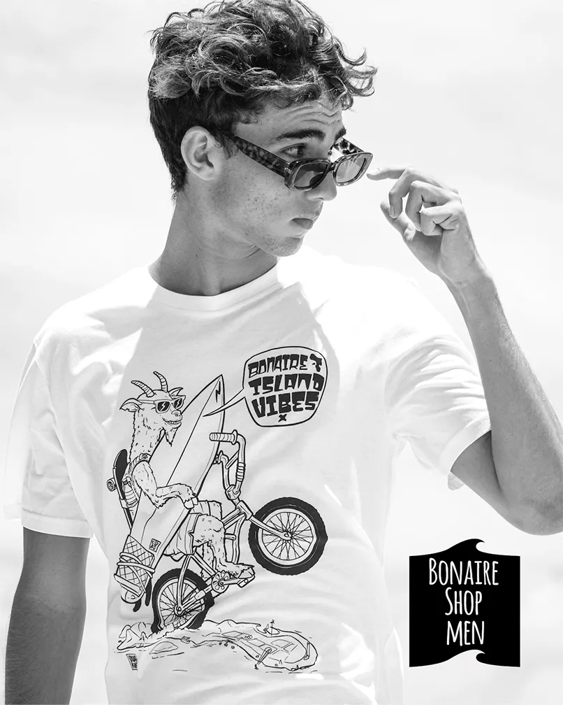 Young man wearing the Bonaire Island Vibe T-shirt with the goat cycling across Bonaire on his back wheel with a surfboard under his arm