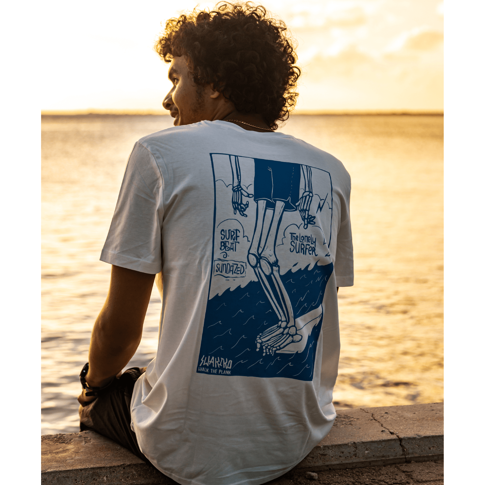 Model aan de kade van Bonaire bij zonsondergang, met een wit T-shirt van de \'eenzame surfer\'