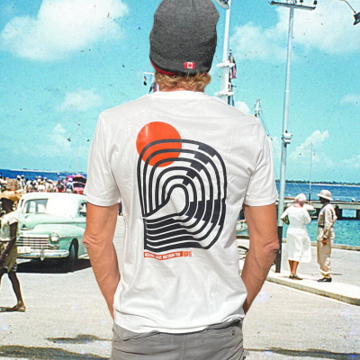 Model staat op de kade van Bonaire en draagt een wit T-shirt met een abstract surf design van een golf en oranje zon