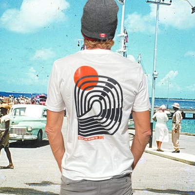 Model staat op de kade van Bonaire en draagt een wit T-shirt met een abstract surf design van een golf en oranje zon