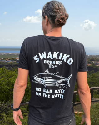 Model draagt een zwart Swakiko T-shirt met een design van tonijn op een surfboard en kijkt uit over de zee van Bonaire