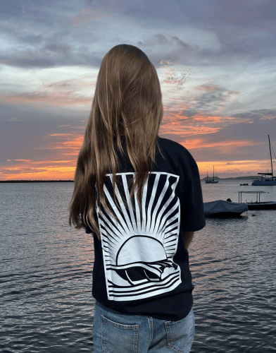 Model staat langs de kade bij zonsondergang met een zwart T-shirt een artistiek design van een golf bij volle maan