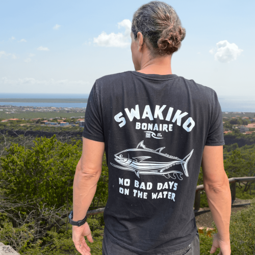 Model draagt een zwart Swakiko T-shirt met een design van tonijn op een surfboard en kijkt uit over de zee van Bonaire
